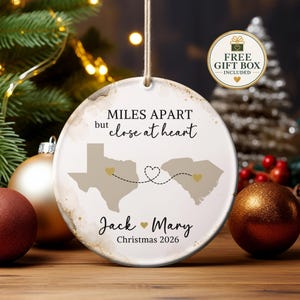 May include: A round ceramic Christmas ornament featuring the text MILES APART but close at heart, with silhouettes of Texas and South Carolina connected by a dashed line and hearts. The names Jack and Mary and the year Christmas 2026 are printed below.