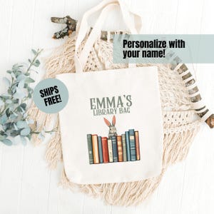 May include: A white canvas tote bag with the text "Emma's Library Bag" and a cartoon rabbit peeking over a stack of colorful books.
