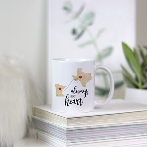 May include: White ceramic coffee mug with a gold outline of Minnesota and Texas with red hearts on each state. The states are connected by a dotted line. The text "always in my heart" is printed on the mug.