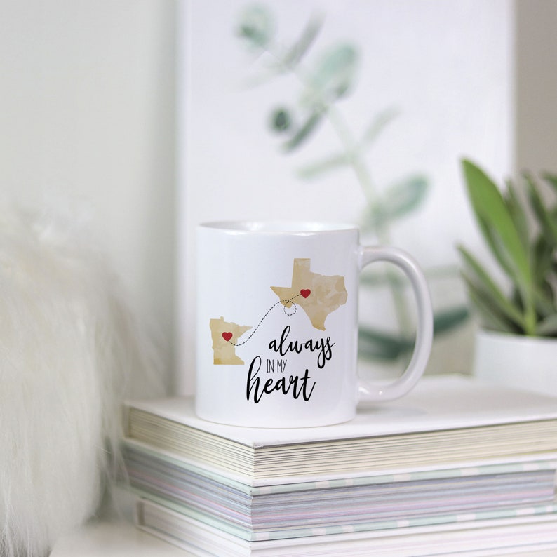 May include: White ceramic coffee mug with a gold outline of Minnesota and Texas with red hearts on each state. The states are connected by a dotted line. The text "always in my heart" is printed on the mug.