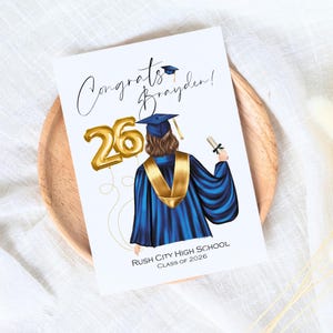 May include: A graduation card featuring an illustration of a graduate in a blue gown and gold stole, holding a diploma. Gold balloon numbers read "26". The card reads "Congrats Brayden!" and "Rush City High School Class of 2026".