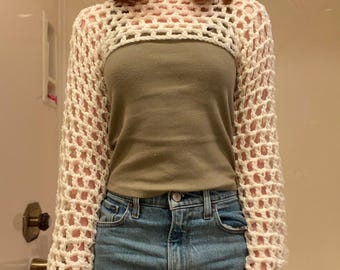 May include: Cream-colored crochet shrug with long sleeves. The open-weave design of the shrug is visible. The shrug is worn over a green top and blue jeans. The style is a cropped bolero.