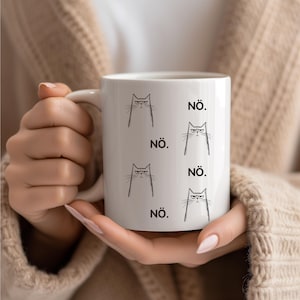 Könnte beinhalten: Weiße Keramik-Tasse mit einem sich wiederholenden Muster aus grimmigen Katzen-Gesichtern und dem Wort "NÖ." in Schwarz. Die Tasse hat einen einfachen Henkel und wird von einer Person gehalten.