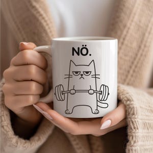 Puede incluir: Taza de cerámica blanca con una ilustración en negro de un gato levantando pesas, con la palabra "NÖ." encima del gato. El gato tiene una expresión enfadada. La taza está sujeta por una persona.