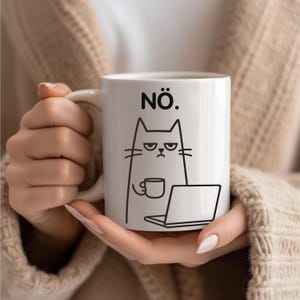 Puede incluir: Taza de cerámica blanca con un dibujo lineal negro de un gato gruñón sosteniendo una taza de café y frente a una computadora portátil. La palabra "NÖ." está impresa encima del gato. La taza es sostenida por una persona.