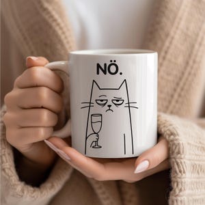 Puede incluir: Taza de cerámica blanca con una ilustración negra de un gato sosteniendo una copa de vino, con la palabra "NÖ." encima del gato. El gato tiene una expresión de mal humor. La taza es sostenida por una persona.