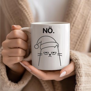 May include: White ceramic mug featuring a grumpy cat wearing a Santa hat. The mug has the word "NÖ." above the cat illustration. The cat has a displeased expression with its eyebrows furrowed. The mug is held by a person.