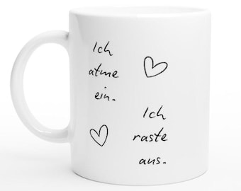 May include: White ceramic mug with a curved handle. The mug features black handwritten text and two heart doodles. The text reads "Ich atme ein. Ich raste aus."