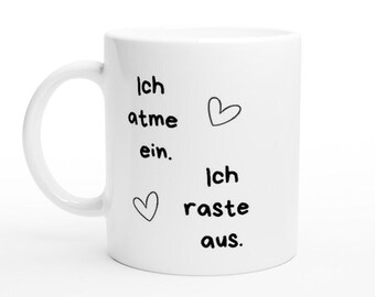 Può includere: Una tazza da caffè in ceramica bianca con un manico curvo. La tazza presenta testo nero e due illustrazioni a forma di cuore. Il testo recita "Ich atme ein. Ich raste aus."