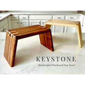 May include: Two handcrafted hardwood step stools. One is a rich brown with lighter wood accents, the other is a light natural wood. Both have angled legs and a rectangular top. The text "KEYSTONE - Handcrafted Hardwood Step Stool -" is visible.