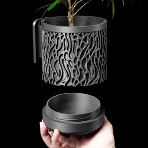 May include: A black, 3D printed planter with a removable lid and a textured, organic pattern. The planter is holding a small plant with green leaves.