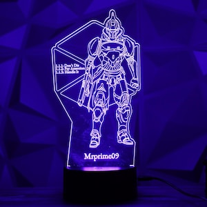 May include: A neon-lit, laser-cut acrylic lamp featuring a detailed outline of a futuristic armored figure. The lamp has the text "Don't Die, Pay Attention, Handle It" and "Mrprime09". The base is black, and the light emits a purple glow.