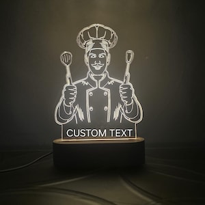 May include: A white acrylic night light featuring a silhouette of a chef holding two kitchen utensils. The base is made of wood and has the text "CUSTOM TEXT" printed on it.