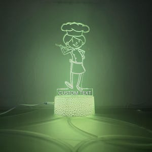 May include: A green night light with a white outline of a chef holding a spatula. The base of the light is textured and has the words "CUSTOM TEXT" printed on it.