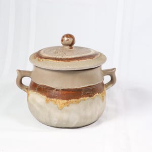 May include: A ceramic pot with a lid, featuring a brown and white glaze. The pot has two handles and a rounded shape.