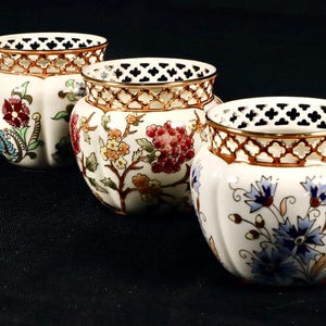 Puede incluir: Tres macetas de cerámica blanca con ribetes dorados y diseños florales intrincados. Las macetas tienen un diseño de celosía alrededor del borde superior.
