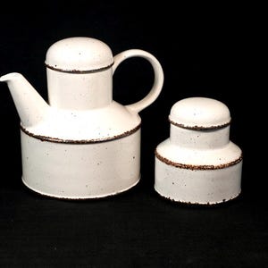 May include: White ceramic teapot and sugar bowl set. The teapot has a curved handle and spout, with a lid. Both pieces have a speckled finish and a brown trim around the base and lid. The set is on a black background.