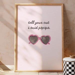 Könnte beinhalten: Ein gerahmter Druck mit dem Text "tell your cat i said pspsps." zeigt eine Aquarellillustration einer rosa herzförmigen Sonnenbrille. Der Druck wird auf einem hellrosa Hintergrund mit einem goldenen Rahmen angezeigt. Darunter befindet sich ein schwarz-weiß karierter Behälter.