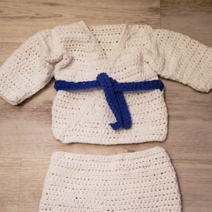 May include: A handmade, white crochet karate uniform set with a blue belt. The set includes a jacket with a V-neck and long sleeves, and matching pants. The outfit is designed for a baby or toddler.