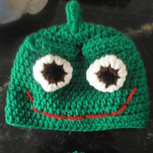 May include: Green crocheted hat with white and brown eyes and a red stitched mouth. The hat is shaped like a frog.