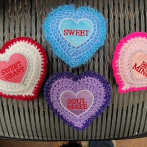 May include: Four handmade crochet heart-shaped decorations. Each heart features a different color scheme: red and white, blue, pink, and purple. Each heart has an embroidered message: "Sweet Heart", "Sweet", "Be Mine", and "Soul Mate".
