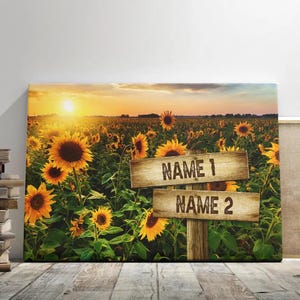 May include: A rustic wooden sign with the text "NAME 1" and "NAME 2" in a field of sunflowers at sunset. The sign is propped up on a wooden post.