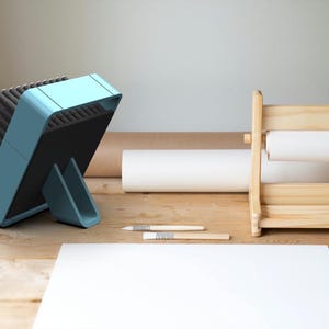 May include: A light blue brush holder with a black base and a stand. The brush holder is filled with a variety of brushes. The brush holder is sitting on a wooden table.
