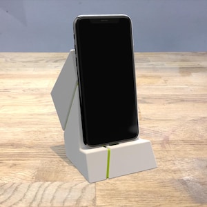 Puede incluir: Un soporte de teléfono blanco con una franja verde, sosteniendo un teléfono inteligente negro en modo vertical.
