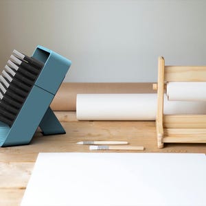 May include: A blue brush holder with a stack of black brushes. The holder is on a wooden surface with a white sheet of paper and a wooden paper roll holder.