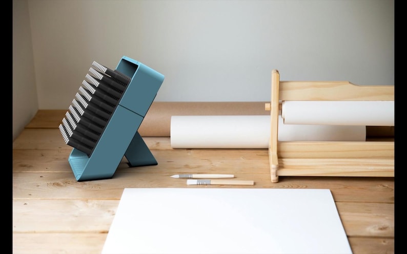 May include: A blue brush holder with a stack of black brushes. The holder is on a wooden surface with a white sheet of paper and a wooden paper roll holder.