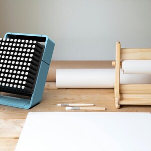 May include: A light blue fan with a black grid of small squares. The fan is sitting on a wooden surface with a white sheet of paper and a wooden paper roll holder.