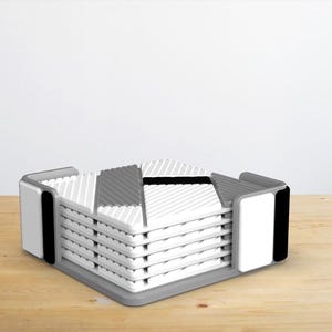 Puede incluir: Un juego de posavasos grises y blancos con un patrón geométrico, apilados en un soporte gris y blanco con detalles negros.
