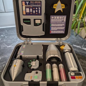 May include: A grey medical kit containing various tools and compartments, including a device with a screen and the text "MEDICAL KIT EQUIPMENT MANUAL." The kit also features a Starfleet insignia and the text "EM MED DG-47."