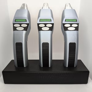 May include: Three silver props with a black base. Each prop features a rectangular green display, two white buttons, and a black vent design. The props are displayed in a black rectangular holder.