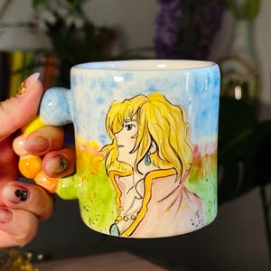 May include: A ceramic mug featuring a hand-painted illustration of a character with blonde hair, set against a backdrop of a blue sky and a field of flowers. The mug has a unique handle with colorful accents.