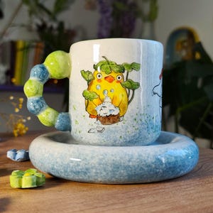 May include: A ceramic mug with a whimsical design featuring a yellow bird with a cupcake, resting on a matching saucer. The handle is a unique, colorful design. The mug and saucer are light blue with a speckled pattern.