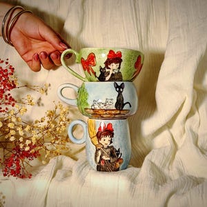 Könnte beinhalten: Drei gestapelte Keramikbecher mit Illustrationen eines Mädchens, einer schwarzen Katze und anderer Katzen. Der obere Becher ist grün mit einer roten Schleife, der mittlere Becher ist blau und der untere Becher ist hellblau. Die Becher sind auf einem weißen Tuch gestapelt.