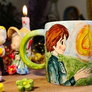 May include: A ceramic mug with a hand-painted illustration of a character with brown hair and a green jacket, facing a yellow flame. The mug has a green handle and is decorated with a landscape scene. Decorative figurines and a lit candle are in the background.