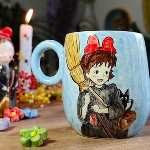 May include: A ceramic mug featuring a cartoon illustration of a girl with a red bow, holding a broom, and a black cat. The mug has a light blue background and a blue handle. Decorative figurines and a lit candle are in the background.