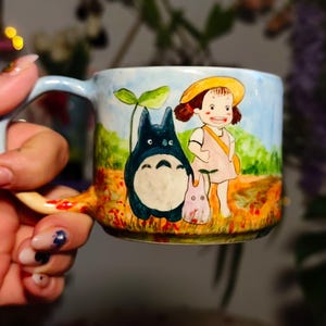 May include: A ceramic mug featuring a hand-painted scene with characters from the animated film "My Neighbor Totoro." The design includes Totoro, a young girl, and a small creature set against a landscape with a blue sky and green grass. The mug has a light blue handle.