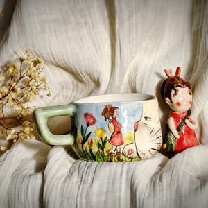 May include: A ceramic mug with a green handle and a painted illustration of a girl in a red dress and a white cat. A small figurine of a girl in a red dress stands beside the mug. The mug is decorated with flowers.