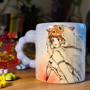 May include: Ceramic mug with a light blue, red, and white design. The mug features a detailed illustration of a character from an animated film, holding a knife. The handle is a unique cloud shape.