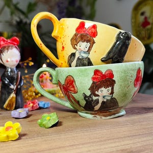 May include: Two ceramic teacups stacked, one yellow and one green, each with a handle. The cups feature illustrations of a girl with a red bow and a black cat. A small figurine and decorative flower-shaped objects are also present.