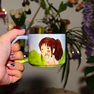 May include: A ceramic mug with a light blue handle and rim, and a green handle base. The mug features a cartoon illustration of a girl with brown hair and a cat. The background is light blue and green.