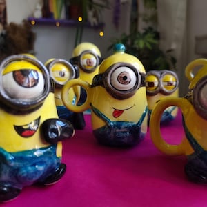 May include: A collection of yellow ceramic Minion-themed teacups and teapots. Each piece features a Minion face with large eyes, black goggles, and blue overalls. Some have handles and spouts, while others are shaped like the Minion characters.