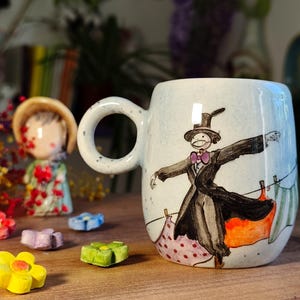 May include: A ceramic mug with a whimsical illustration of a character in a black coat and top hat, with a pink bow tie. The mug has a large, round handle and is decorated with a light blue speckled background. Colorful ceramic flowers and figurines are in the background.