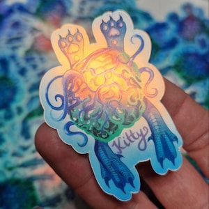 Op de afbeelding: Een holografische sticker met een schildpadachtig wezen met een gloeiend, veelkleurig hoofd en blauwe ledematen. De sticker heeft het woord "Kitty" in een scriptletter.