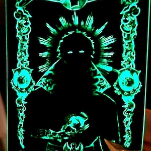 Può includere: Una carta verde che brilla al buio con una silhouette nera di una figura con un alone di luce. La figura tiene un piccolo oggetto nella mano. La carta è circondata da un bordo decorativo con teschi e catene.