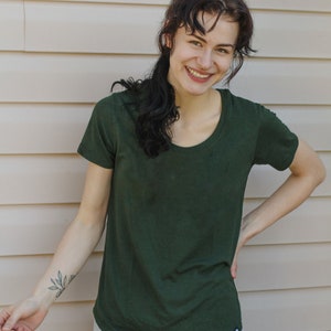 May include: A woman wearing a dark green t-shirt with a small black logo on the bottom right corner. She is smiling and has dark hair. She is wearing light blue jeans.