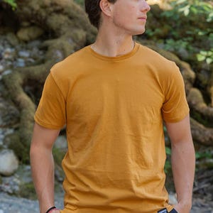 May include: A mustard yellow short-sleeved crew neck t-shirt with a small black rectangular label on the bottom left hem featuring the white text Kindred Coast. The shirt is made of a solid-coloured fabric and has a standard fit.